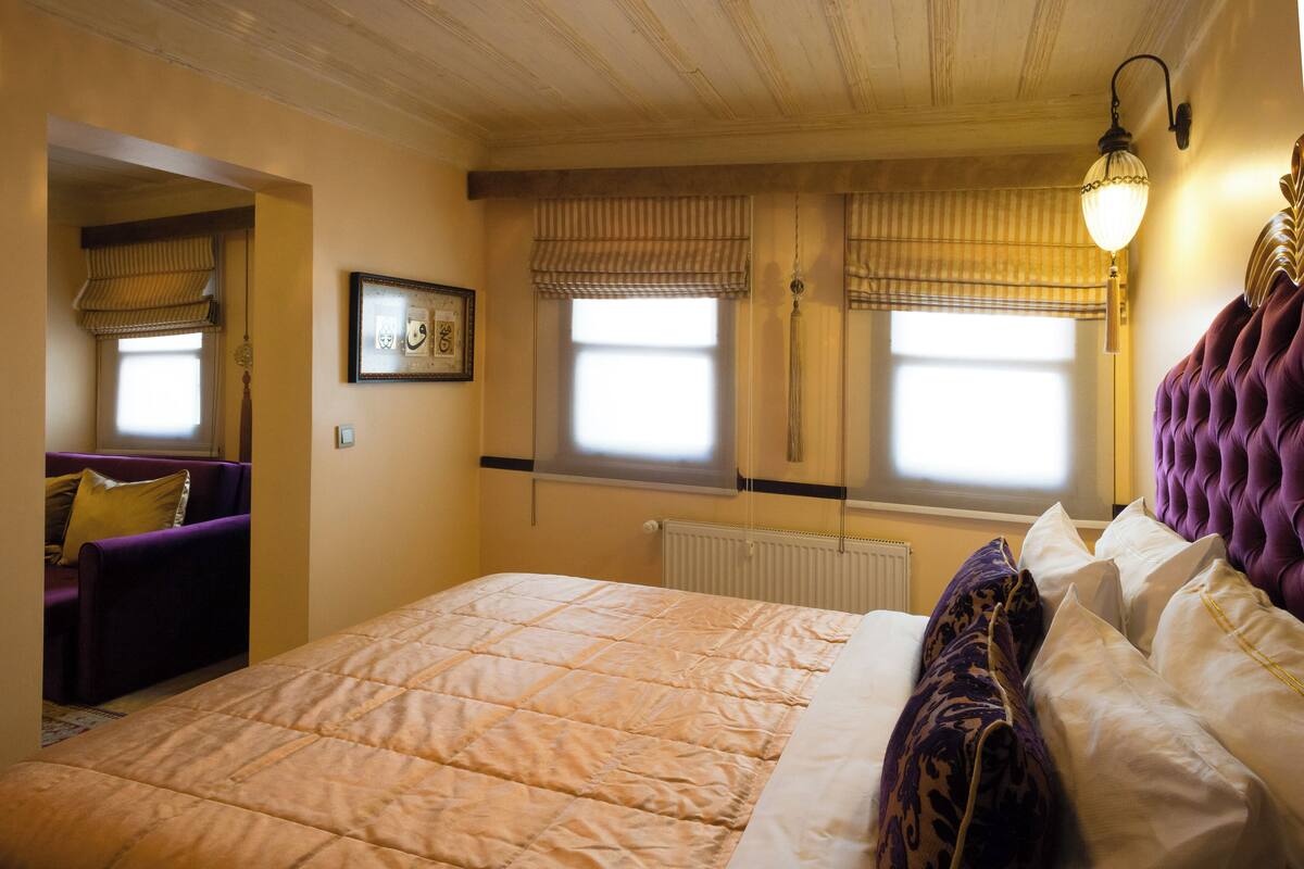 Quarto com uma cama grande, duas janelas com persianas, um radiador, um quadro na parede e um sofá roxo no canto.