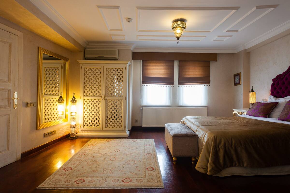 Quarto com cama, mesa de cabeceira, luminária, janela com persianas e porta com espelho e iluminação.