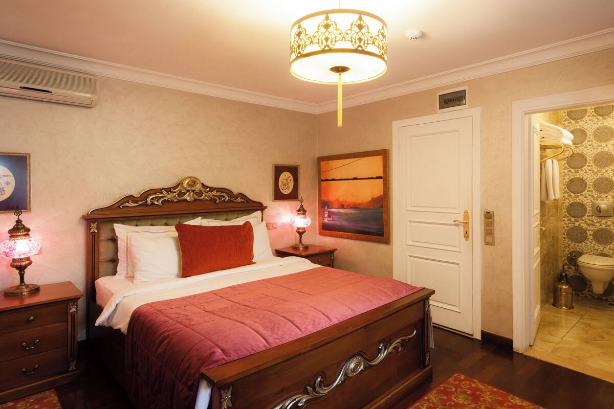 Quarto com cama de madeira, mesa de cabeceira com abajur, pintura na parede e banheiro visível por uma porta aberta.
