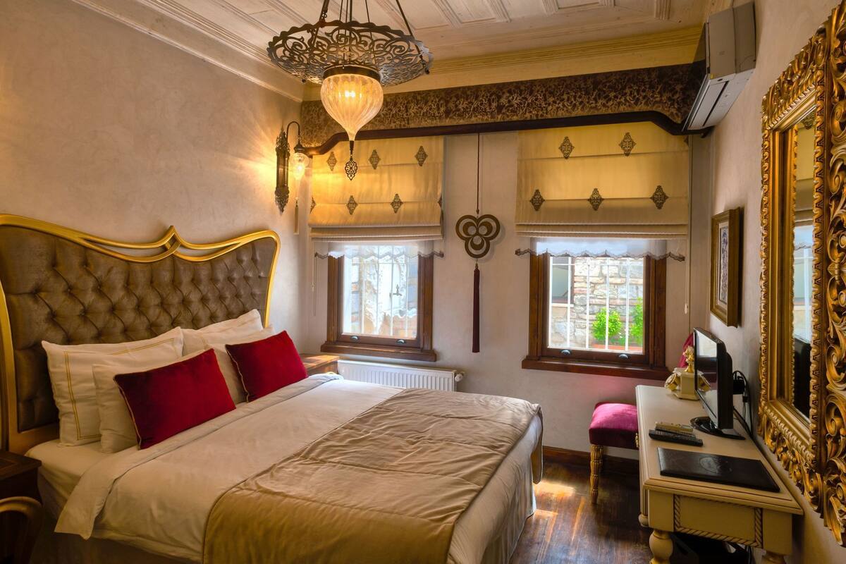 Quarto com uma cama grande, um candelabro, uma escrivaninha com um computador e um espelho grande e ornamentado.