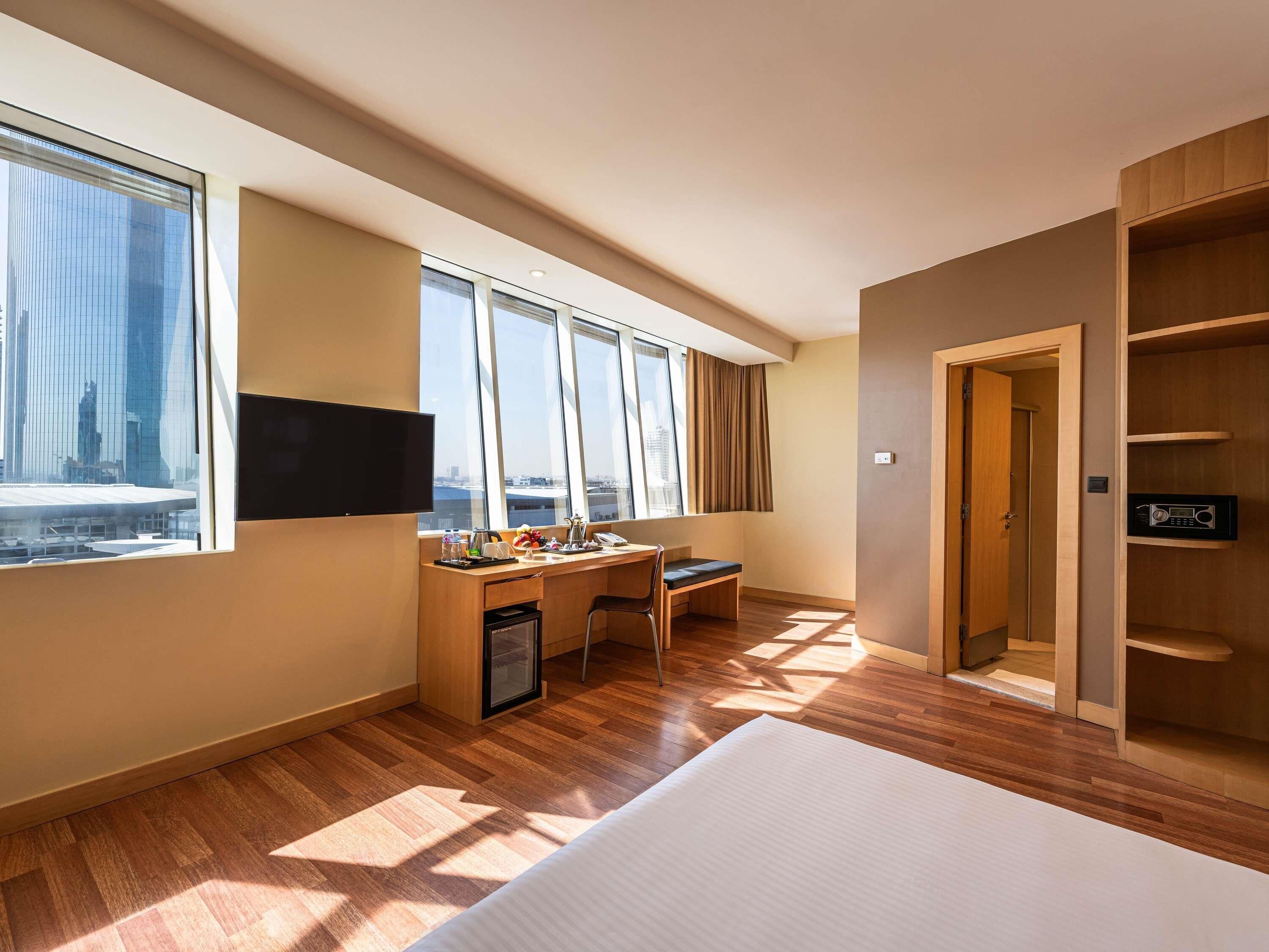 Une chambre d’hôtel avec un grand lit, un bureau avec une chaise, une télévision et une vue sur le paysage urbain grâce à de grandes fenêtres.