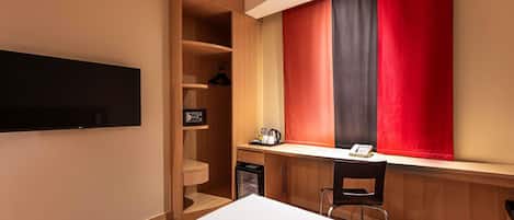 A hotel room with a bed, a desk with a chair, a TV mounted on the wall, and a wardrobe with a mirror.