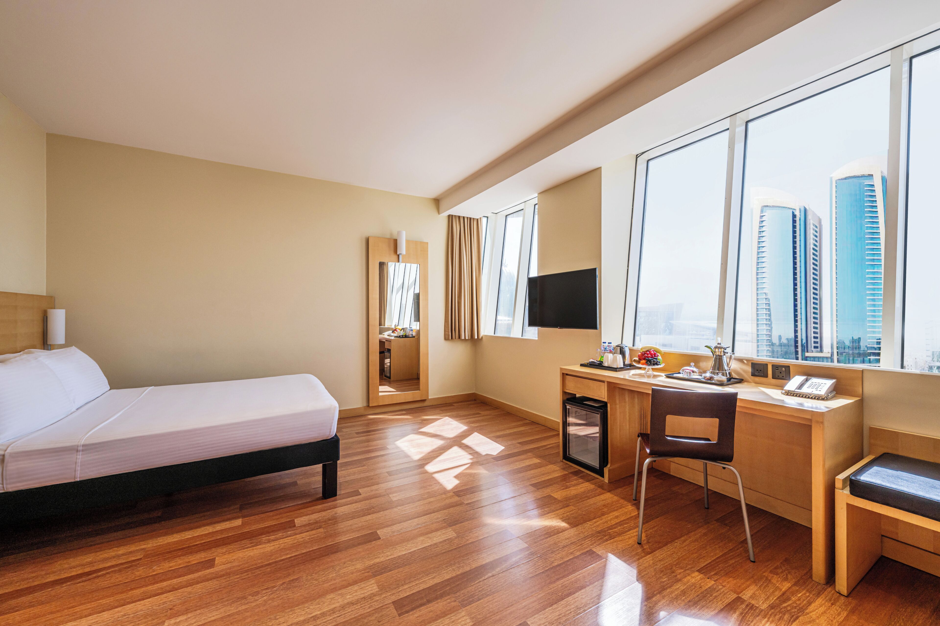 A hotel room with a bed, desk, chair, and a large window with a city view.