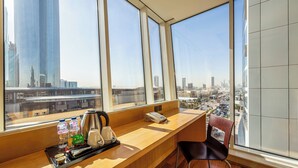 A hotel room with a large window offering a city view, a wooden desk with a tray of amenities, and a chair.