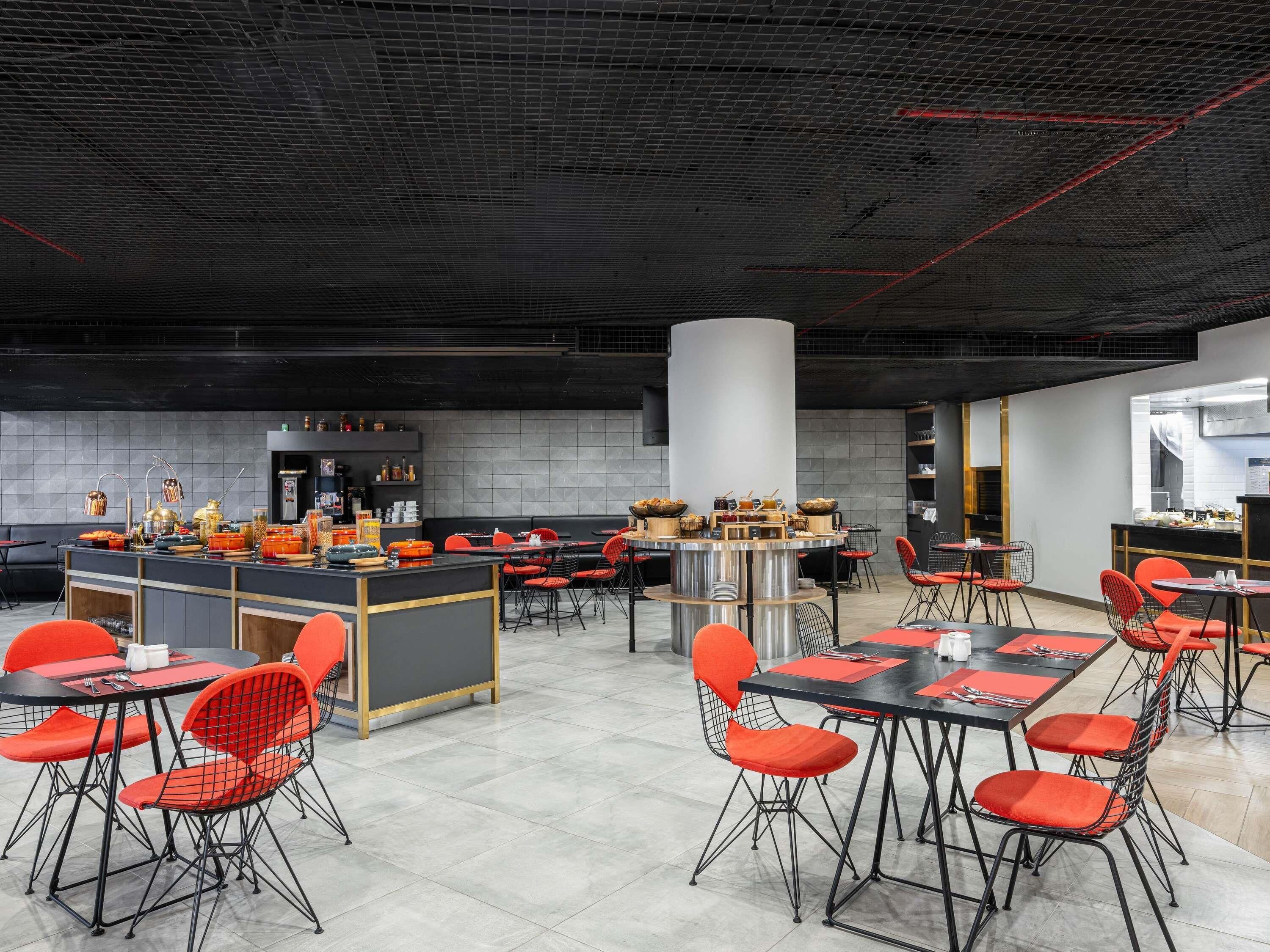 En modern restaurang med en central bardisk, röda stolar och ett kaklat golv.
