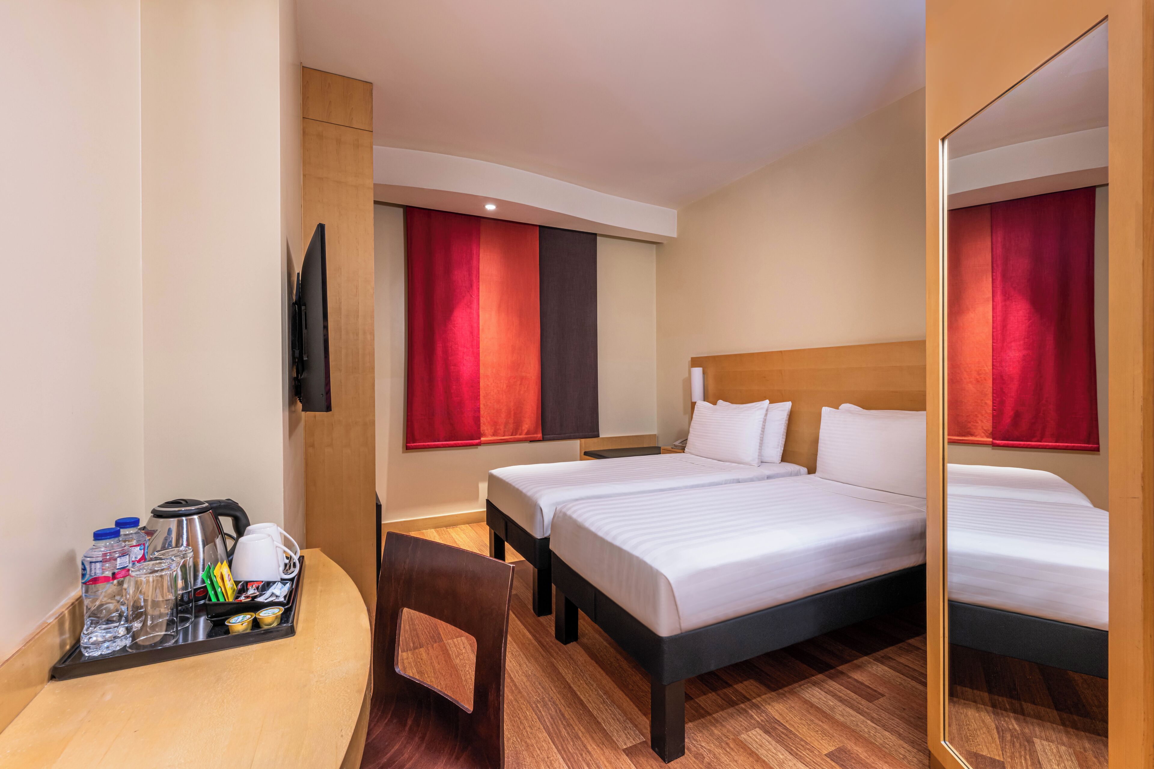 Ein Hotelzimmer mit zwei Betten, einem Schreibtisch mit einem Tablett mit Annehmlichkeiten, einem Sessel und einem Fernseher.