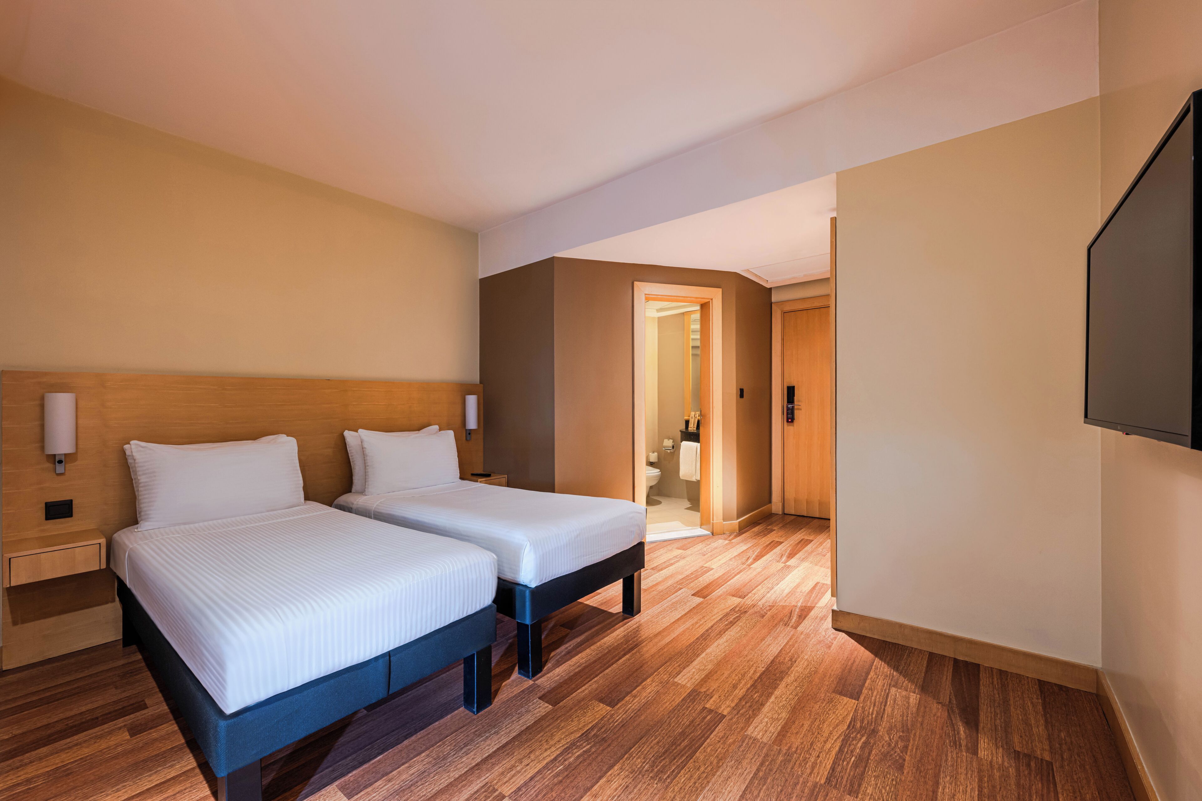 Habitación de hotel con dos camas, suelo de madera, televisión y baño visible a través de una puerta abierta.