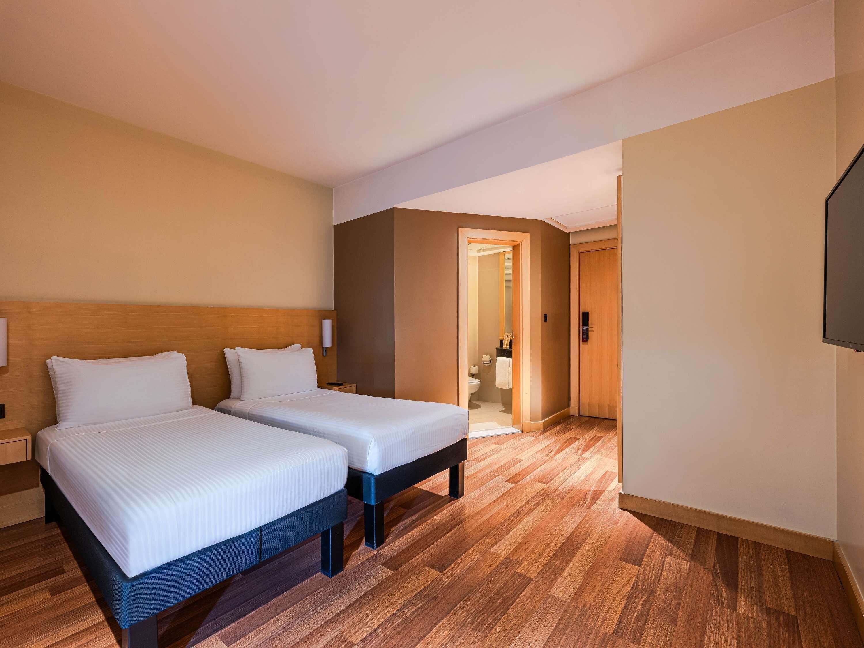 Habitación de hotel con dos camas, cabecero de madera, mesita de noche, televisor de pantalla plana y piso de madera.