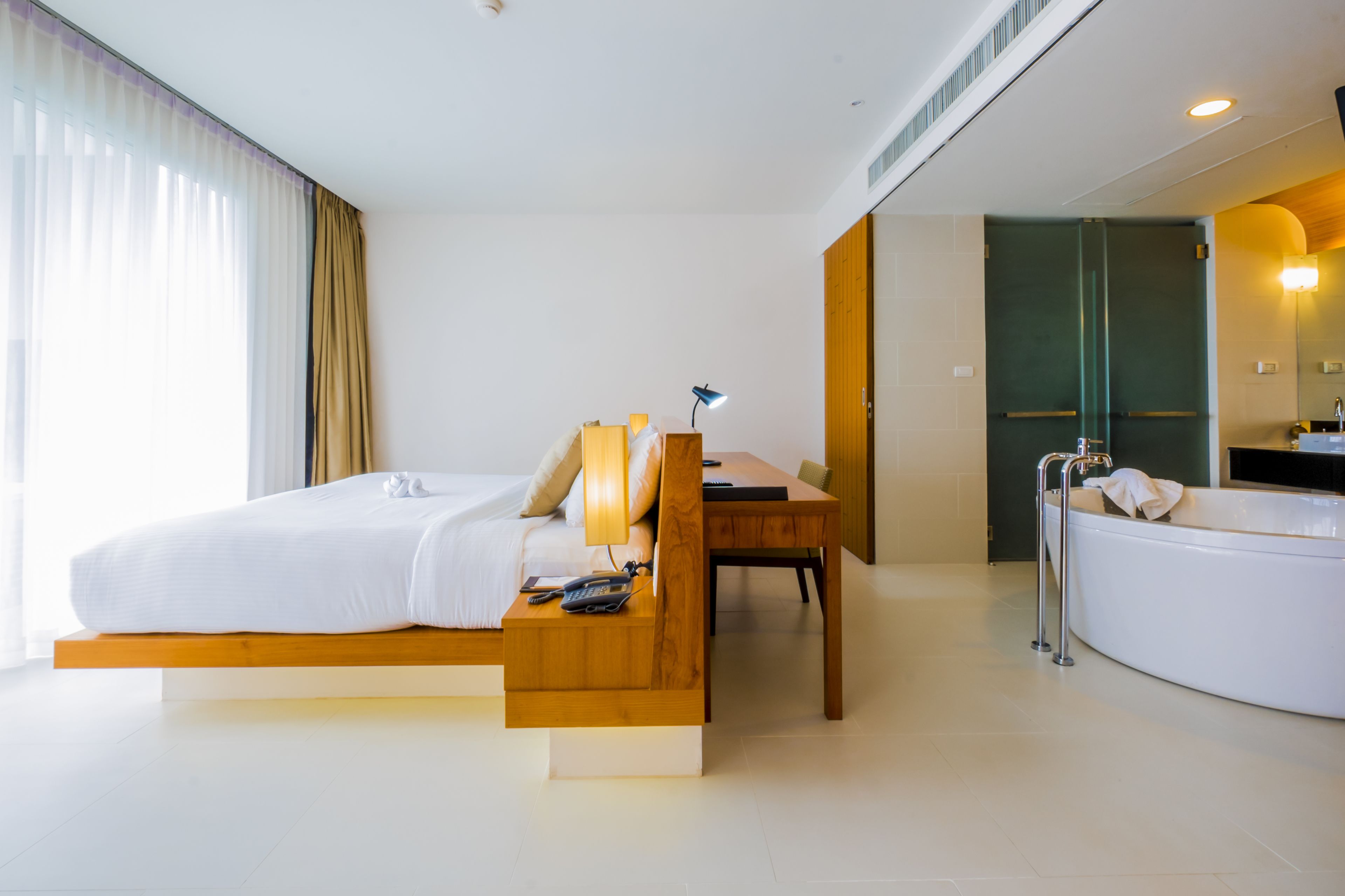 Una habitación de hotel moderna con cama, un escritorio de madera, una lámpara de escritorio, teléfono y bañera.