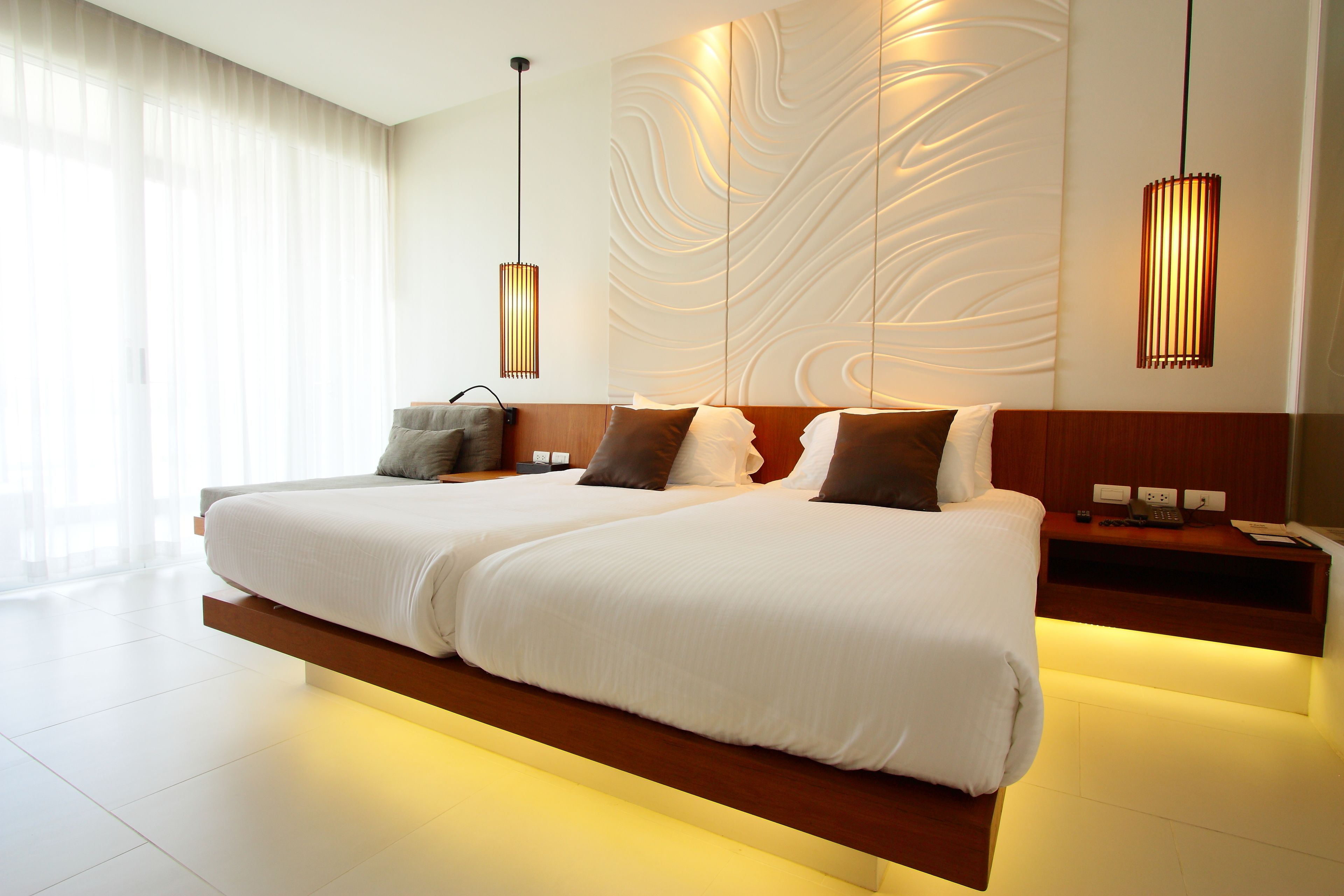 A modern hotel room with two beds, a wooden headboard, a wall with a textured design, and pendant lighting.