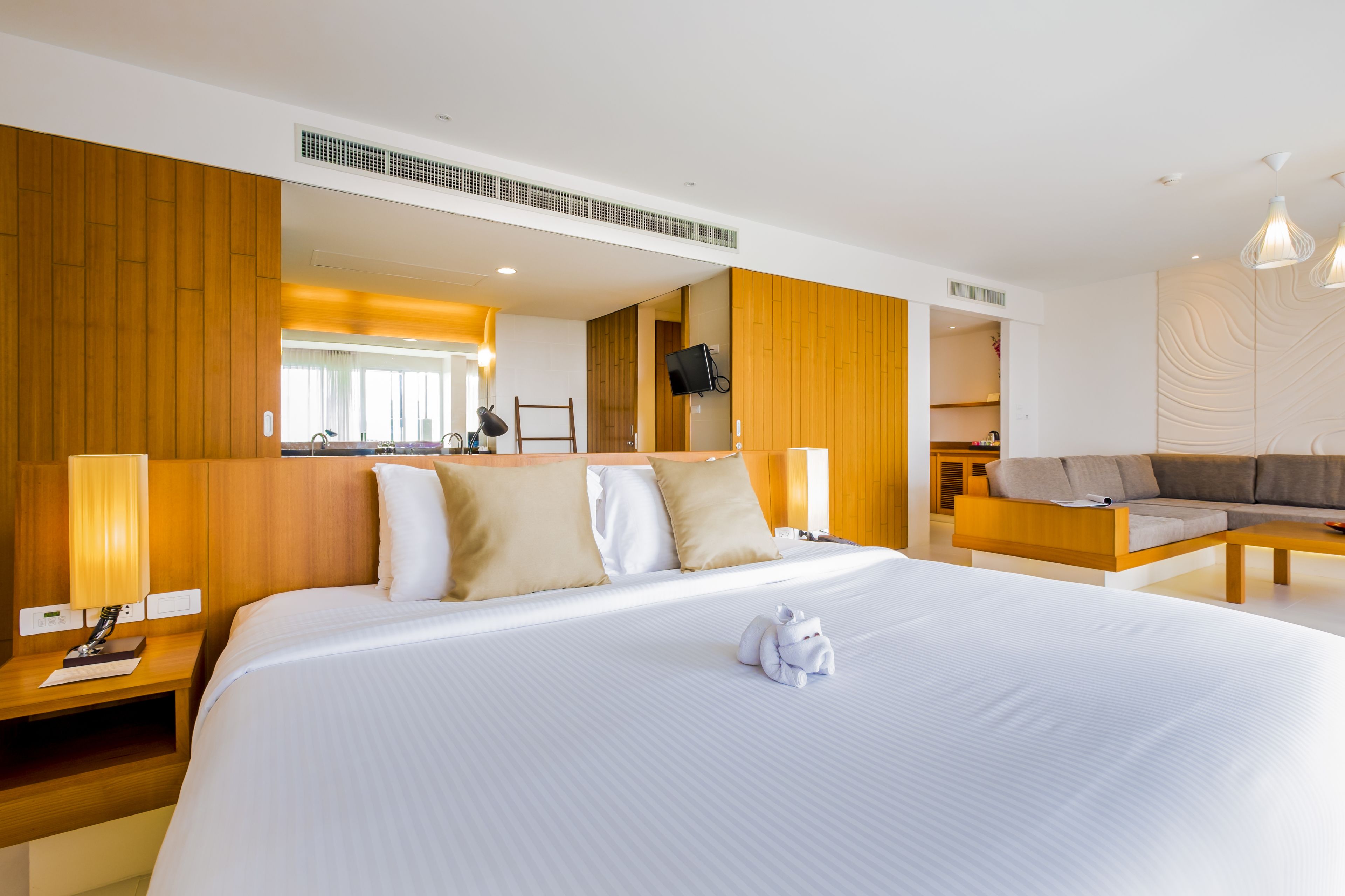A hotel room with a large bed, wooden headboard, bedside tables, a flat-screen TV, and a sofa in the living area.