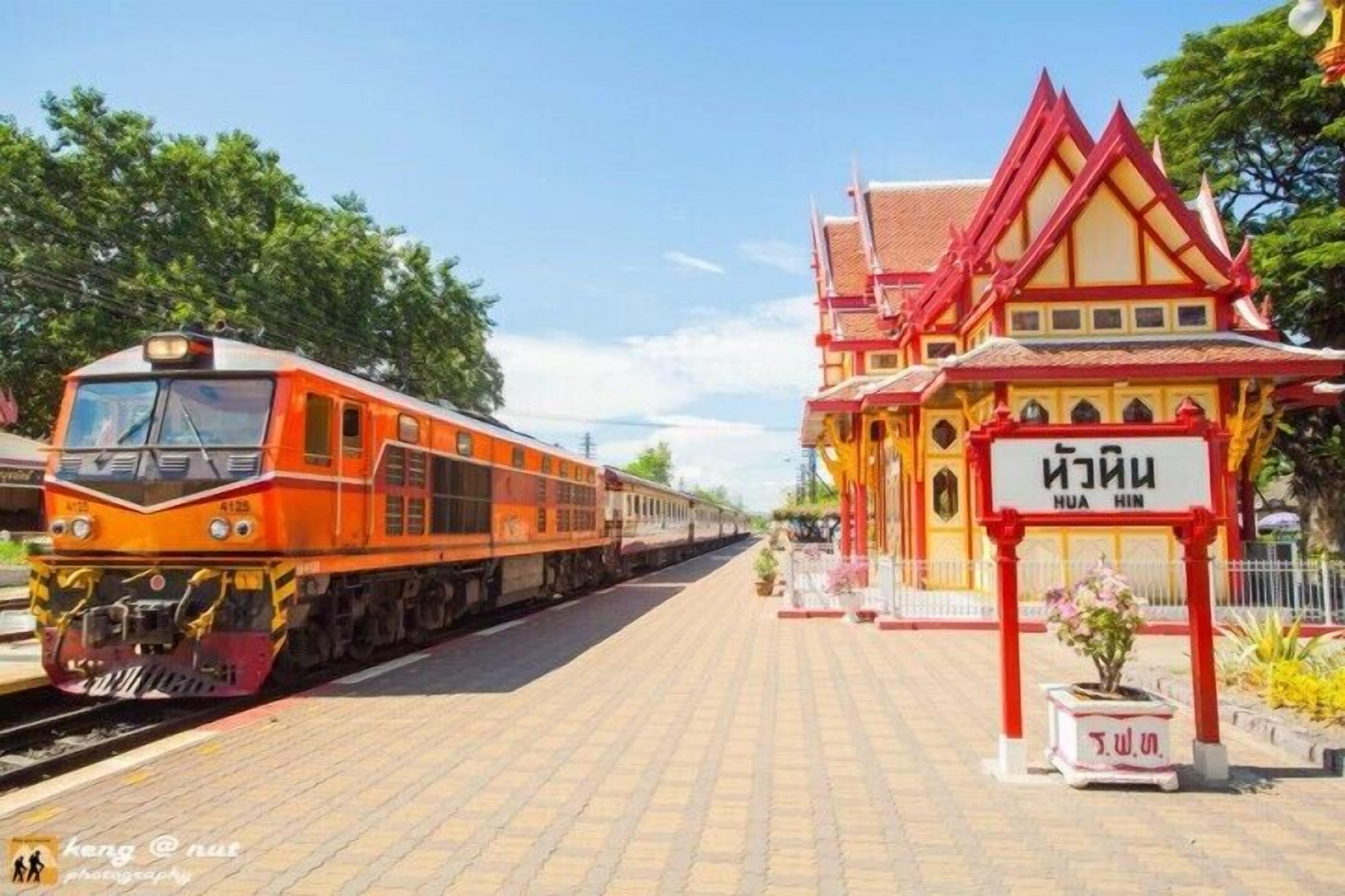 Un andén de estación de tren con un tren rojo y amarillo, un edificio tradicional con un letrero, y un cielo despejado.