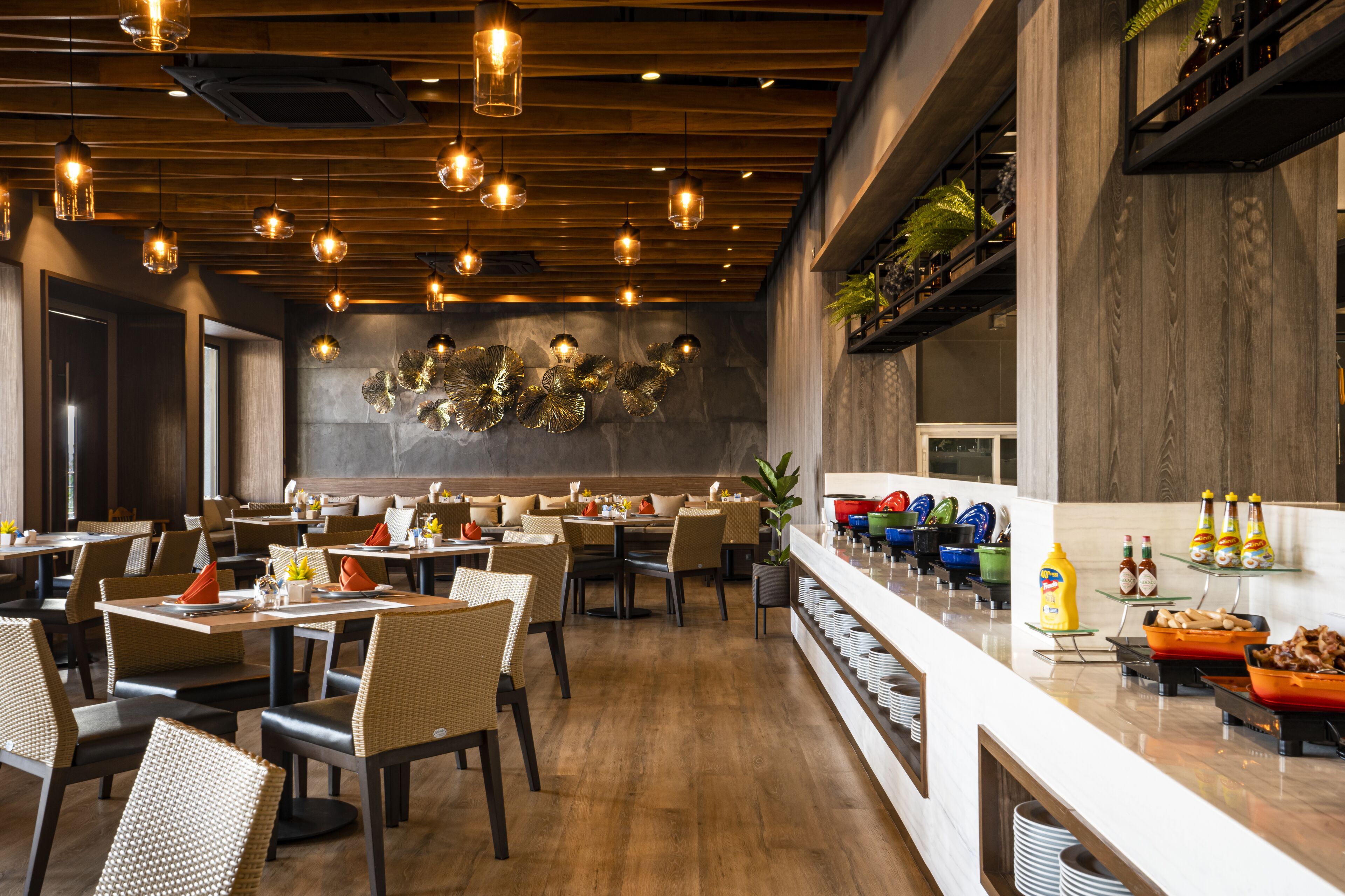 A modern restaurant with wooden ceiling and hanging lights, a buffet with various food items, and a stone wall with decorative elements.