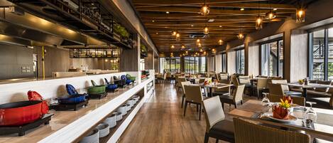 A modern restaurant interior with wooden floors, long communal tables, and hanging pendant lights.