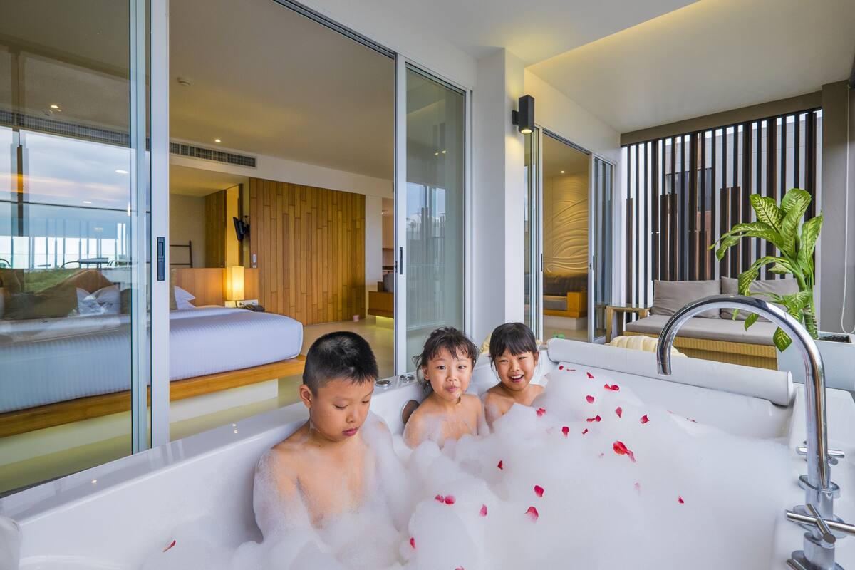 Une chambre d’hôtel moderne avec une grande baignoire remplie de mousse et de pétales de rose, un lit avec du linge de maison blanc et une vue sur l’extérieur.