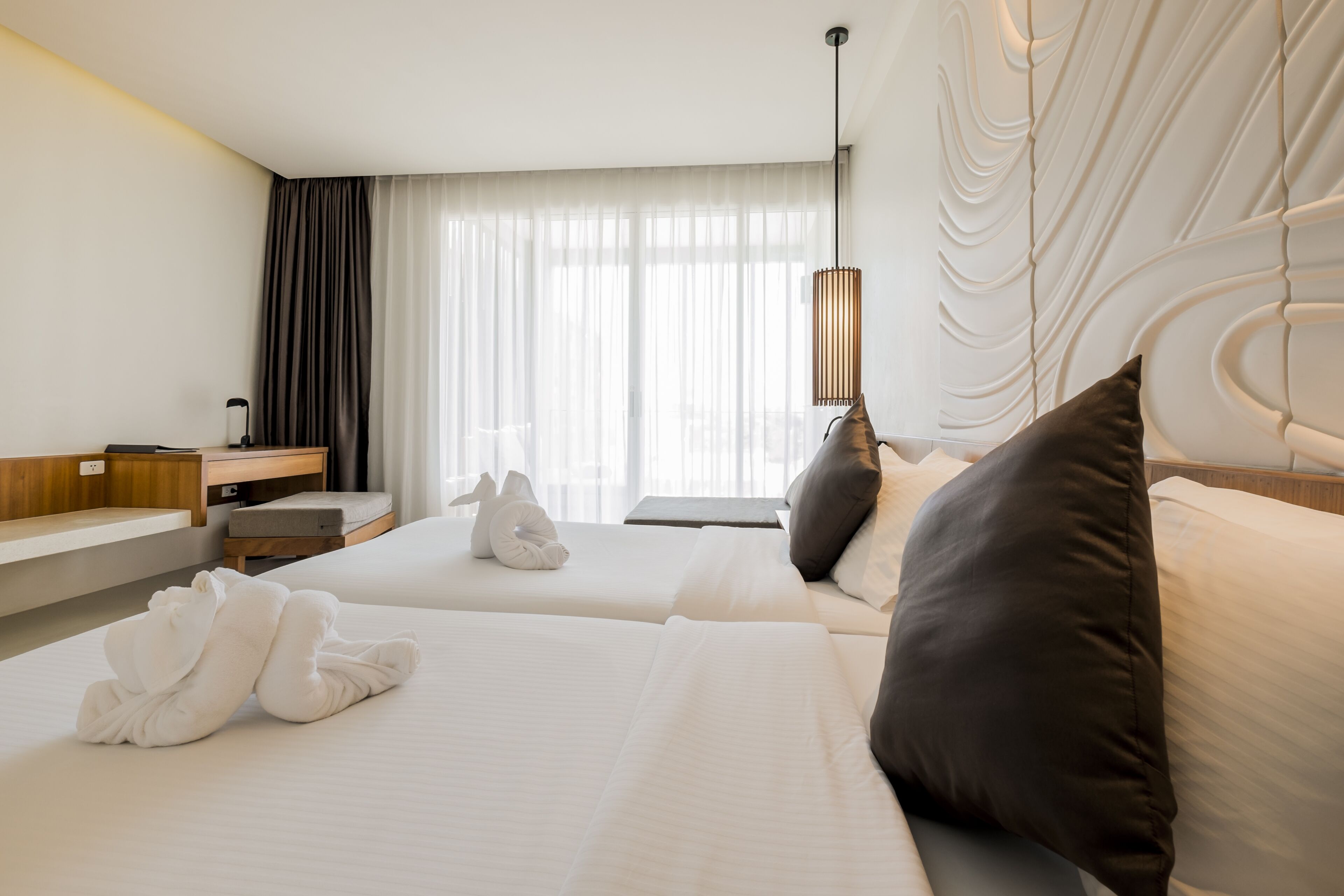 A hotel room with two beds, a desk, a chair, a lamp, and a window with curtains.