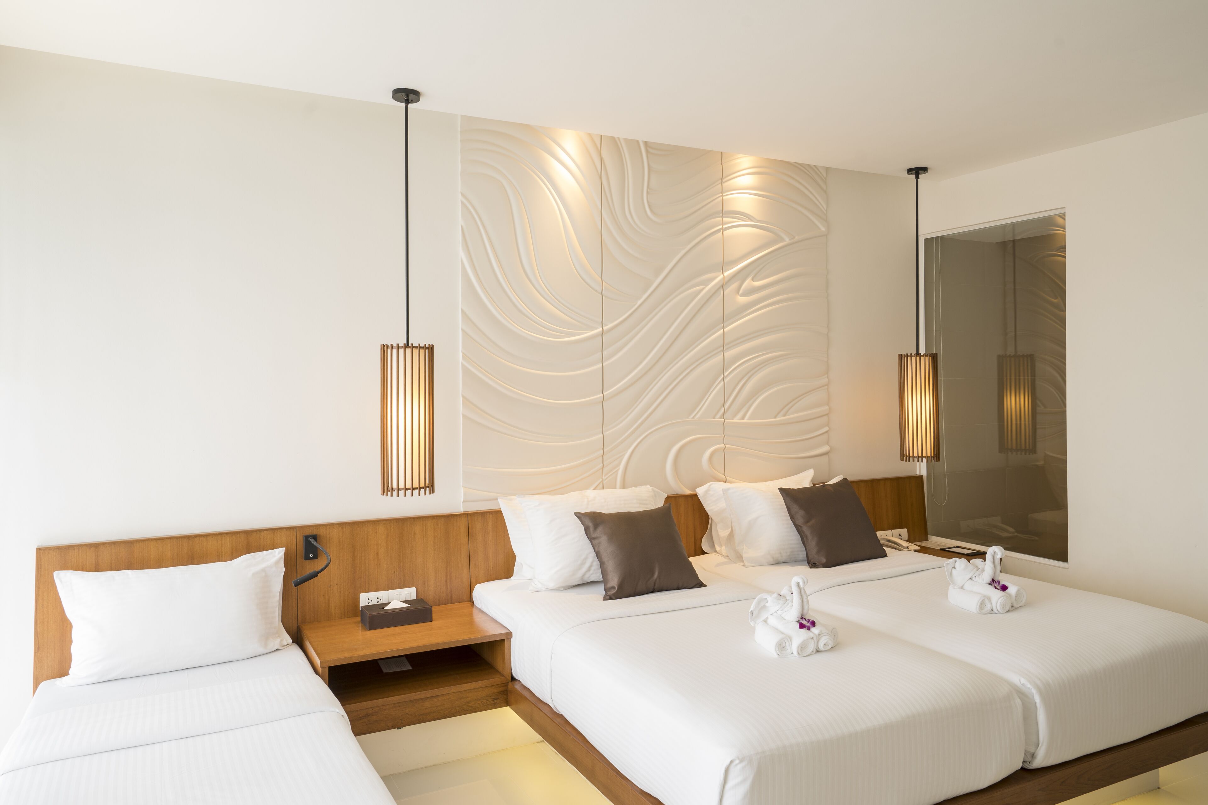 A hotel room with two beds, a wooden headboard, a bedside table with a lamp, and a wall-mounted light fixture.