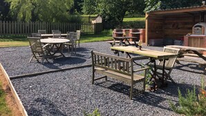 An outdoor patio area with a wooden table and chairs, a barbecue grill, and a wooden structure with a chimney. - Burton Farmhouse (Kingsbridge)