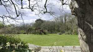 A park with a wooden play structure, a grassy area, and a tree in the foreground. - Burton Farmhouse (Kingsbridge)