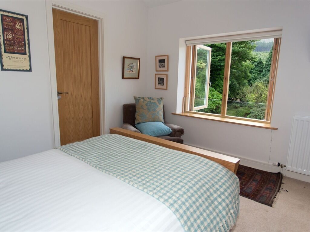 Un dormitorio con cama, ventana que da a zonas verdes, puerta de madera y una pequeña mesa con silla.