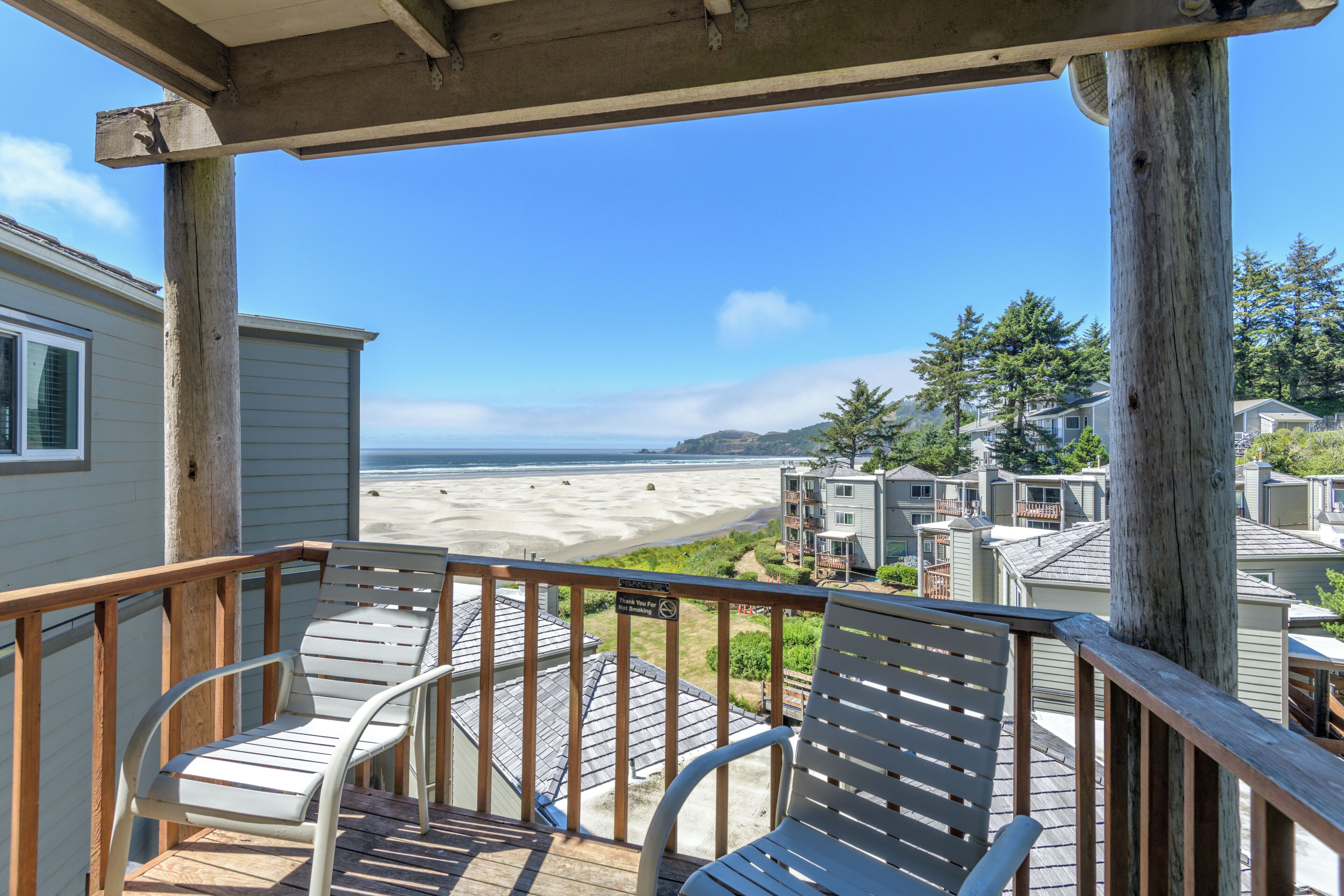 A wooden deck with two white lounge chairs, overlooking a beach and residential buildings.