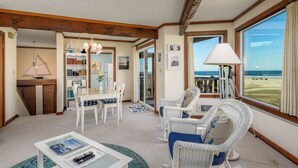 A spacious room with a dining area, a seating area with wicker chairs, a bookshelf, and a large window offering a view of the beach.