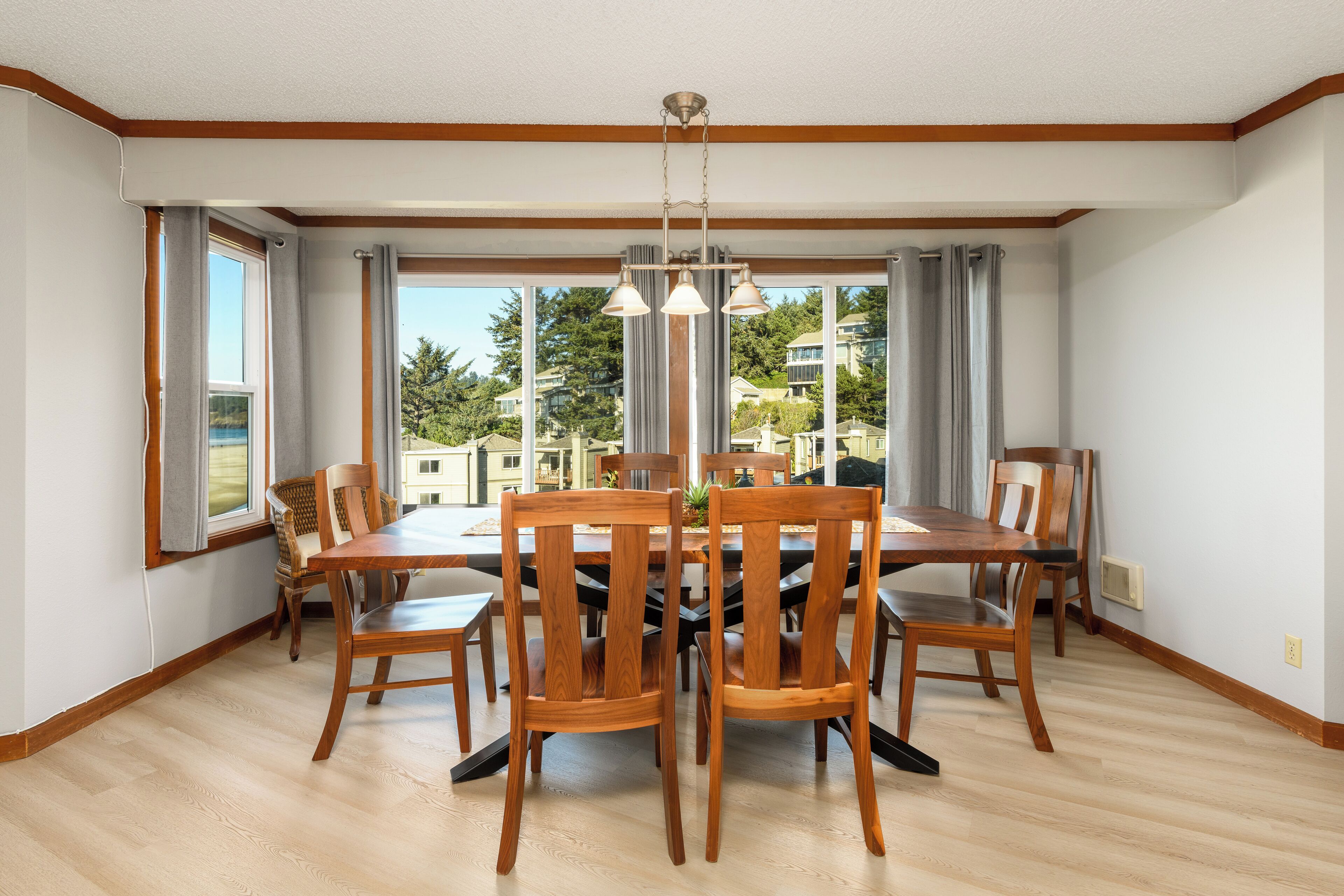 Un comedor con una mesa y sillas de madera, ventanas grandes con cortinas y vista a una zona residencial.