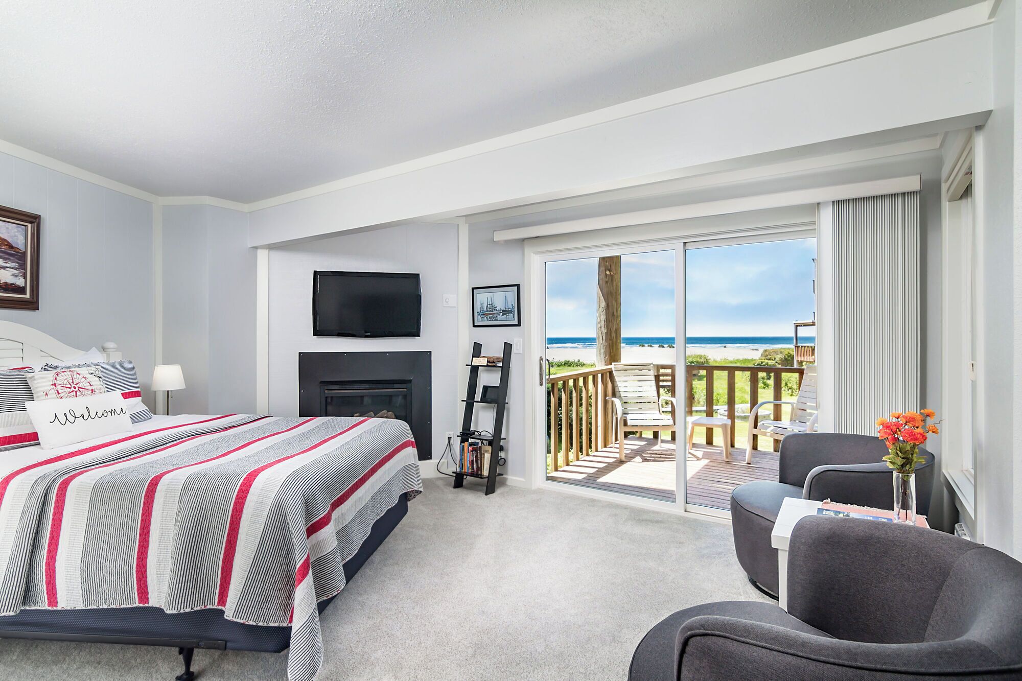 A bedroom with a bed, a TV, a fireplace, and a balcony with a view of the beach.