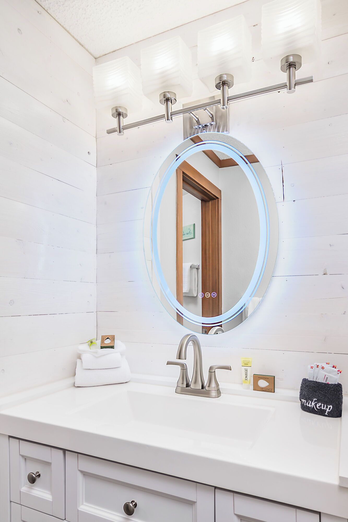 Een badkamer met een wit wastafelmeubel, een ronde spiegel met LED-verlichting, een zilveren kraan en een make-up tas op het aanrecht.