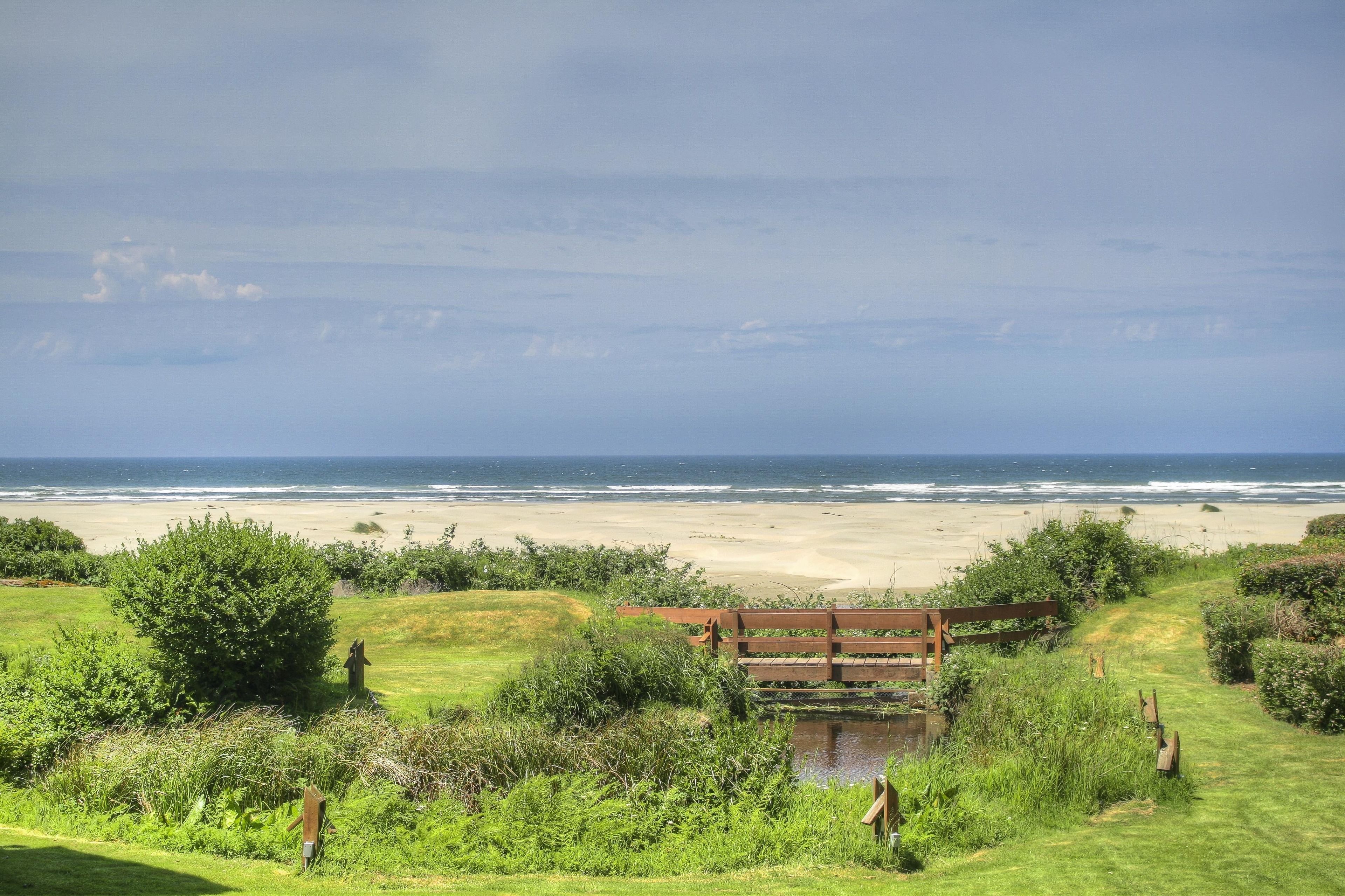 A coastal landscape with a grassy area, a wooden bridge, and a view of the beach and ocean.