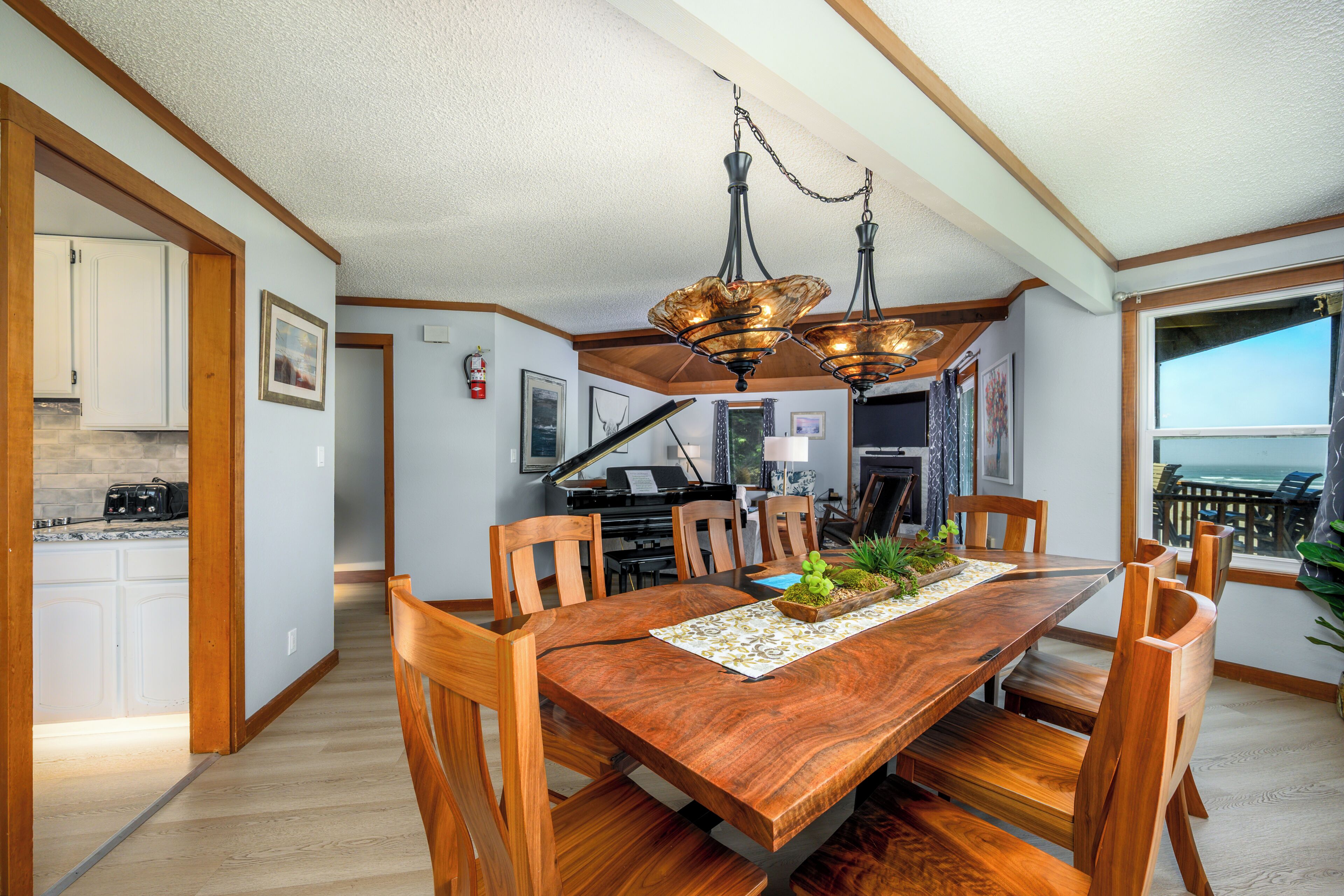 A dining area with a wooden table, chairs, and a chandelier. There is a piano in the background, and a view of the ocean through a window.
