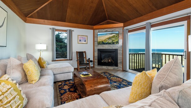 A spacious living room with a fireplace, a sofa set, a wooden coffee table, and a large window offering a view of the ocean.
