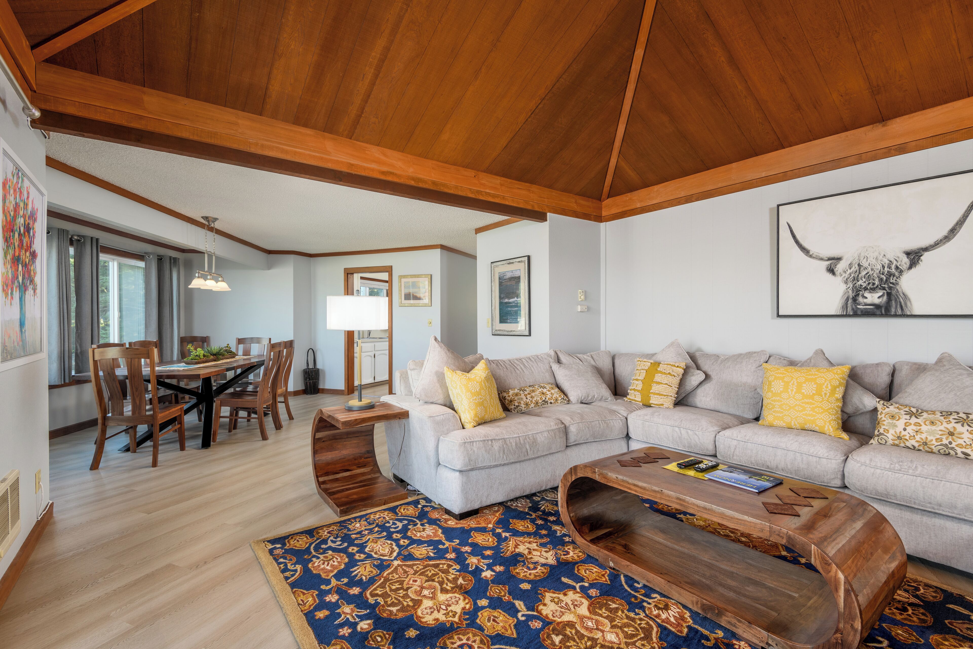 A living room with a wooden ceiling, a gray sofa with yellow pillows, a patterned rug, and a dining area with wooden furniture.