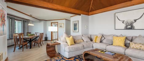 A living room with a wooden ceiling, a gray sofa with yellow pillows, a patterned rug, and a dining area with wooden furniture.