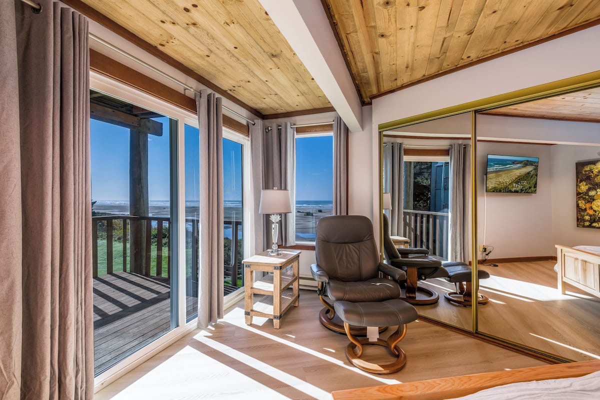 Una habitación amplia con ventanas grandes, techo de madera, sillón de cuero, una mesita con lámpara y una cama.