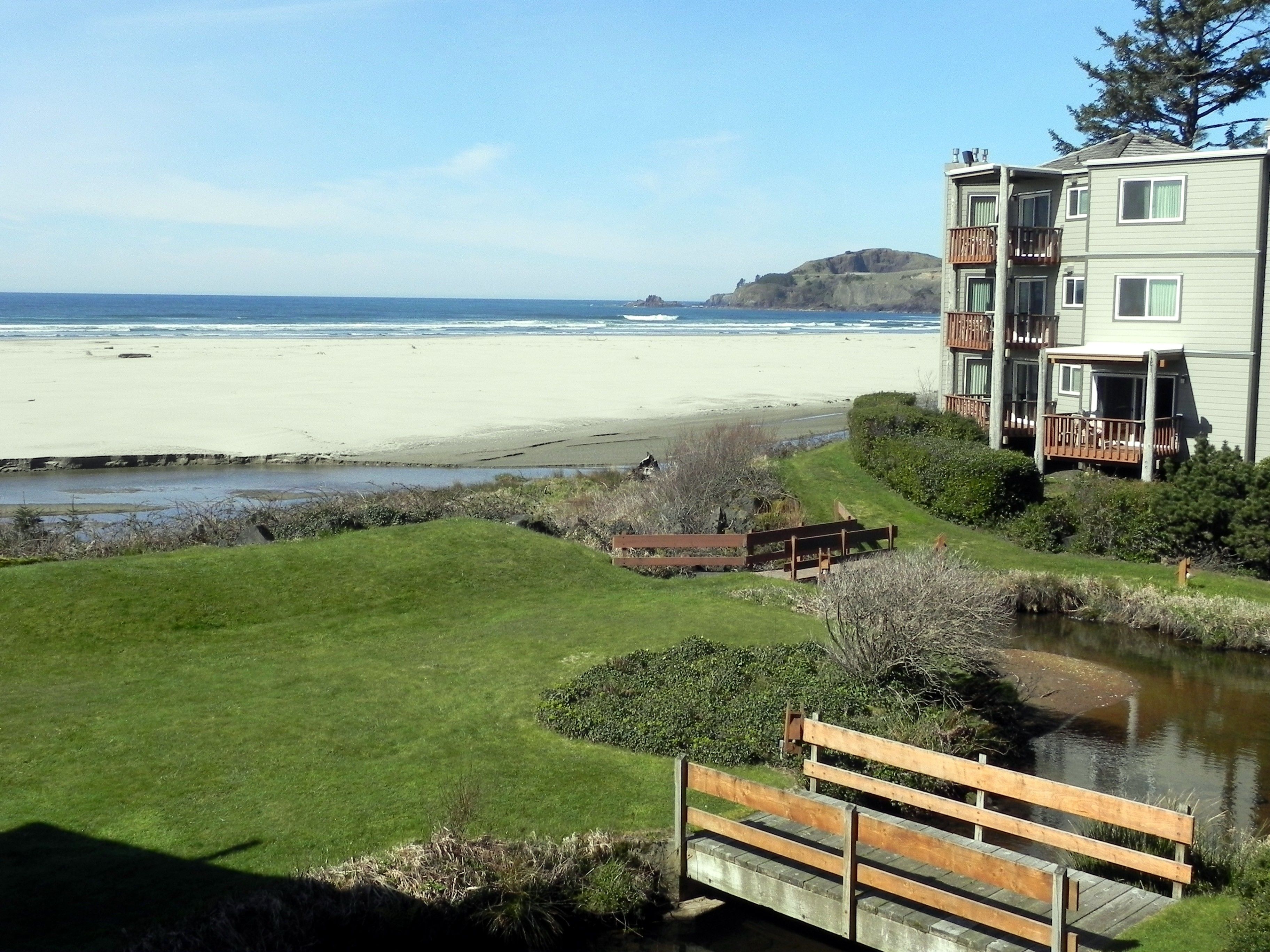 A view of a beach with a grassy area, a wooden bridge, and a building with balconies overlooking the ocean.