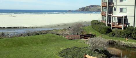 A view of a beach with a grassy area, a wooden bridge, and a building with balconies overlooking the ocean.
