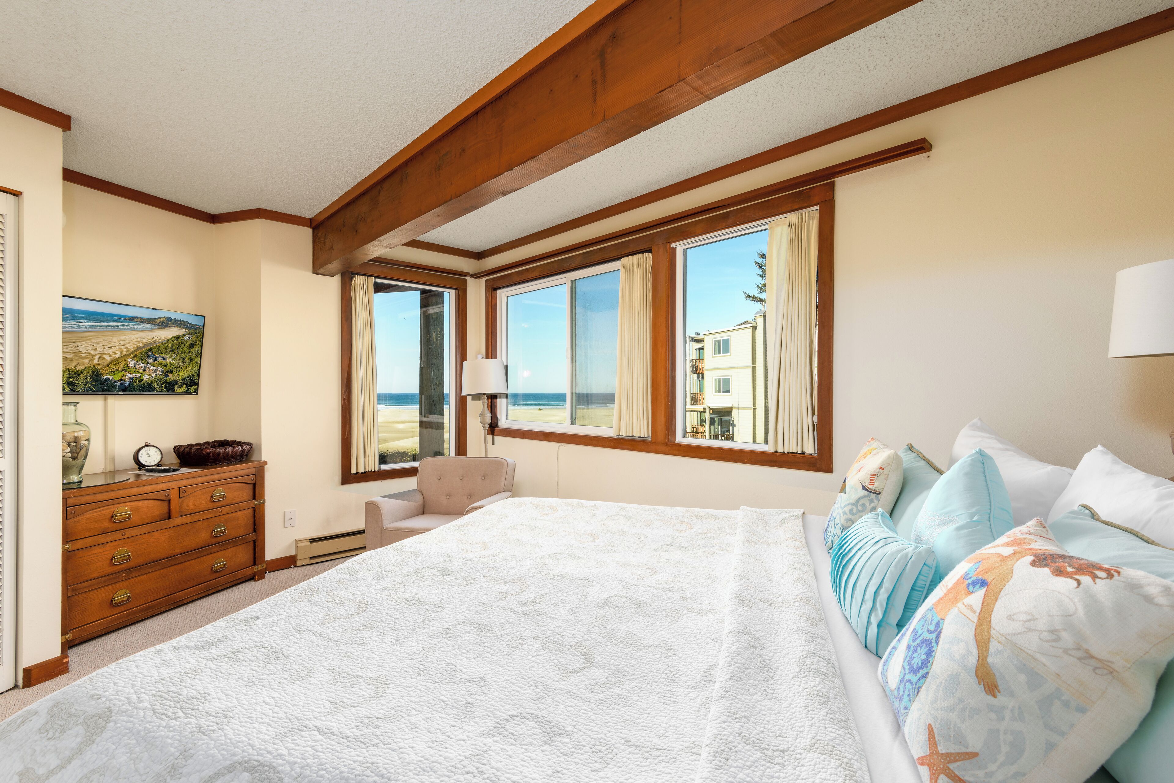 Un dormitorio con una cama grande, vigas de madera, un aparador y vistas a la playa desde las ventanas.