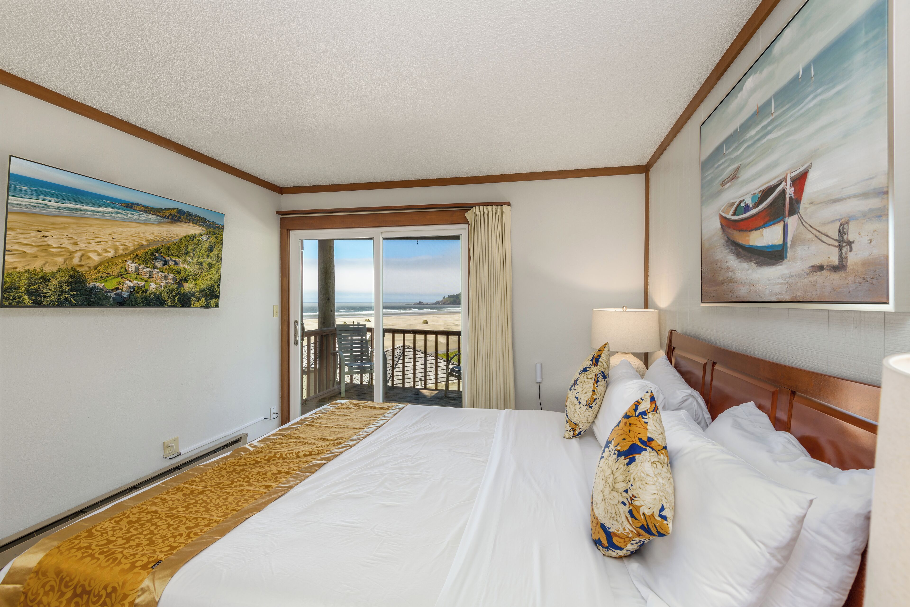 A hotel room with a large bed, a balcony with a view of the beach, and a painting of a boat on the wall.
