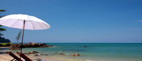 A beach with a wooden lounge chair and a white umbrella, a sandy shore, and clear blue water.