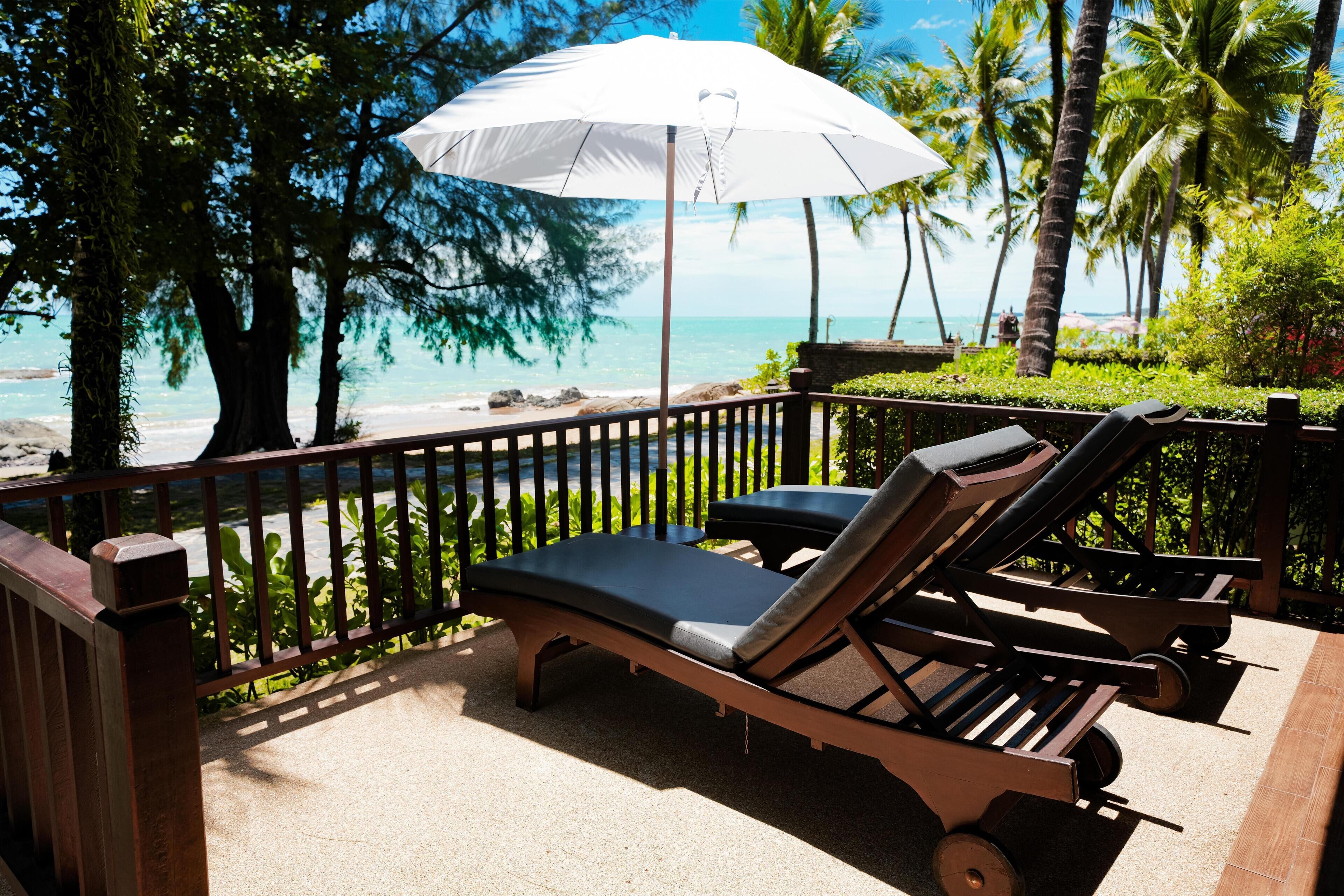 A sun lounger with a white umbrella, a wooden deck, and a view of the beach and ocean.