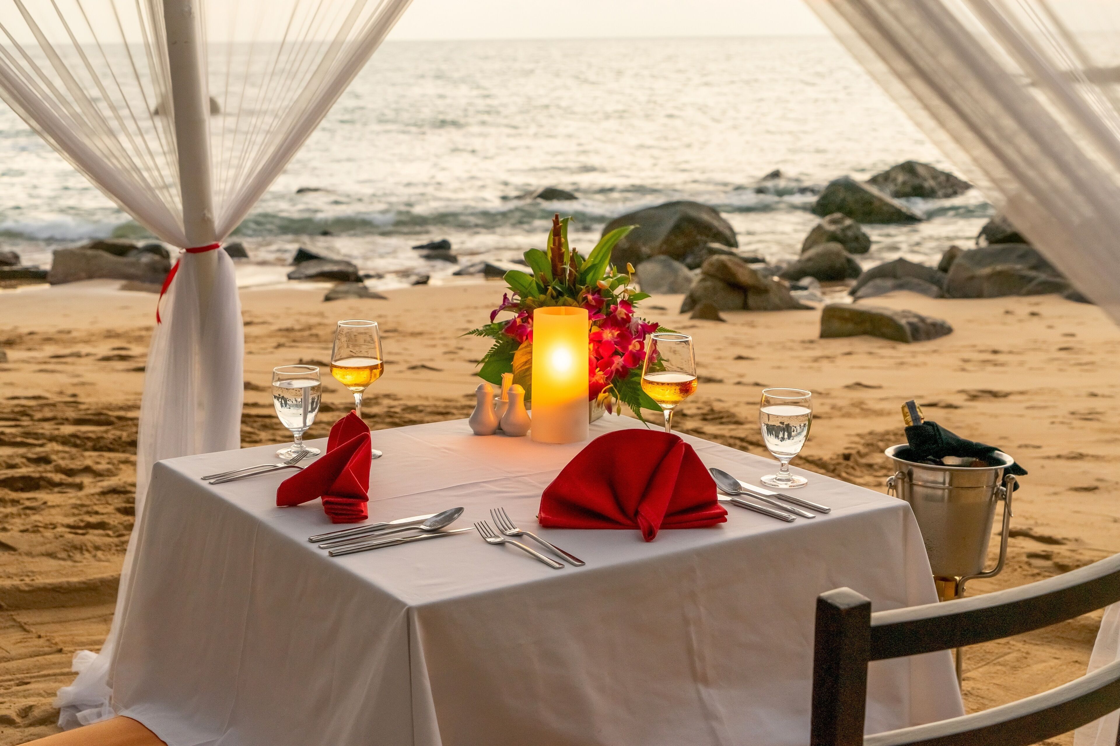 A table set for two with a candle centerpiece, glasses, and a bottle of champagne on a beach.