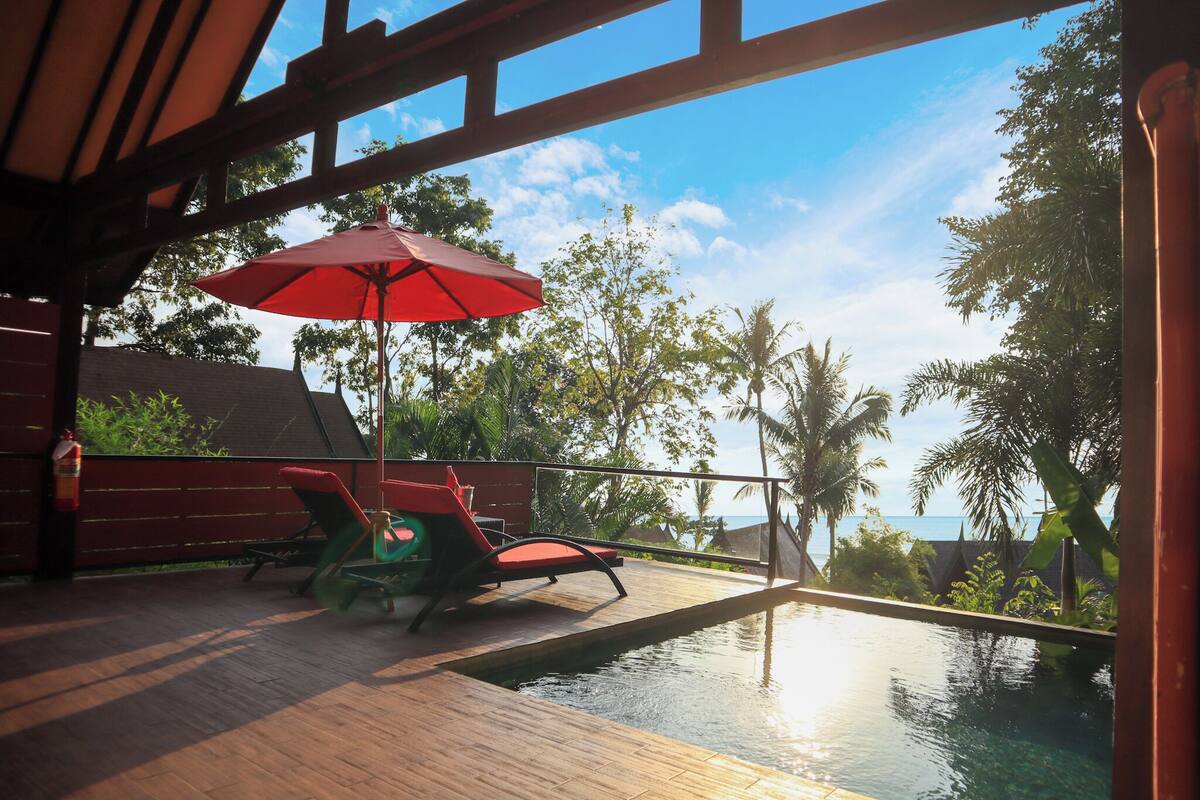An infinity pool with a wooden deck, red lounge chairs, and a red umbrella.