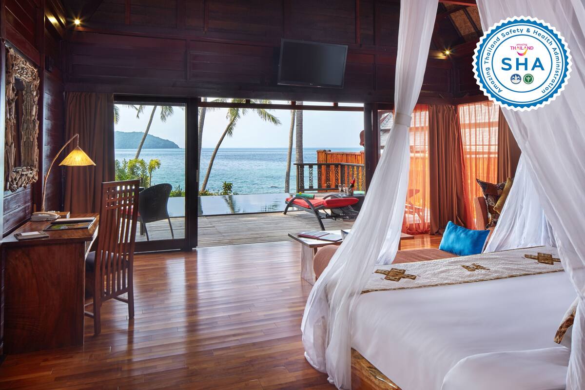 A bedroom with a bed, a desk, and a chair. There is a view of the ocean and palm trees.