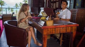 A wooden desk with a laptop, a telephone, and a tissue box in a room with a view of palm trees and the ocean.