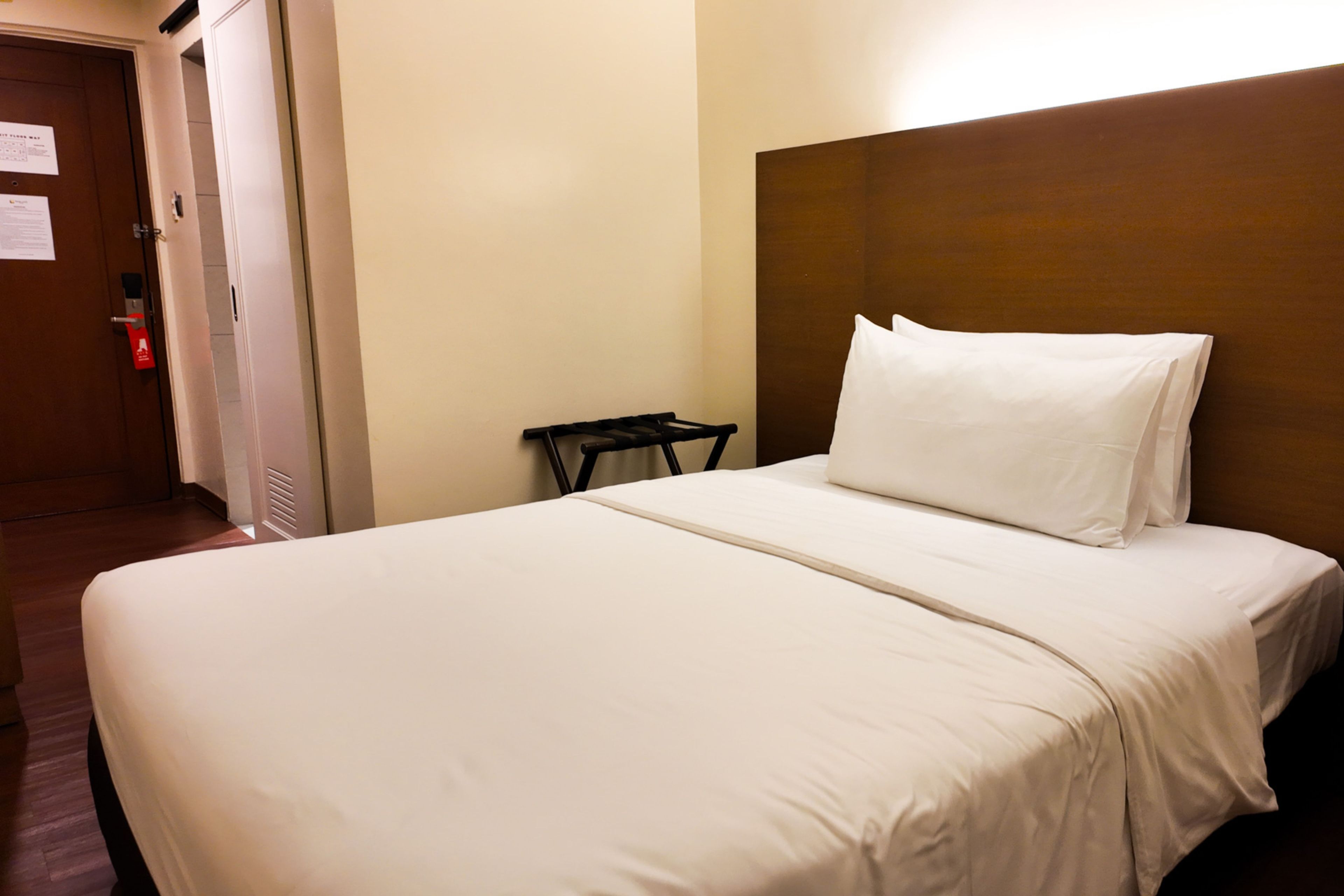 A hotel room with a large bed, a nightstand, a wall-mounted headboard, and a door with a fire alarm.