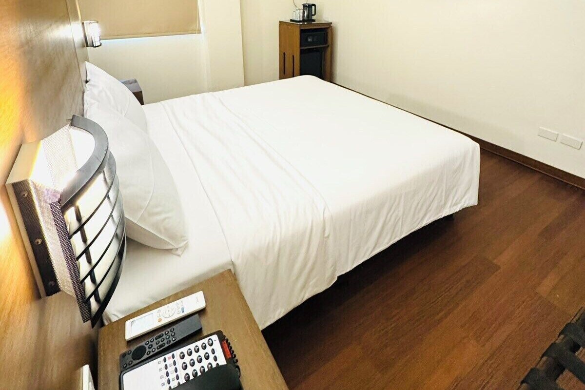 Ein Hotelzimmer mit einem Bett, einem Nachttisch mit Telefon, einem an der Wand befestigten Fernseher und einem kleinen Schrank mit Klimaanlage.