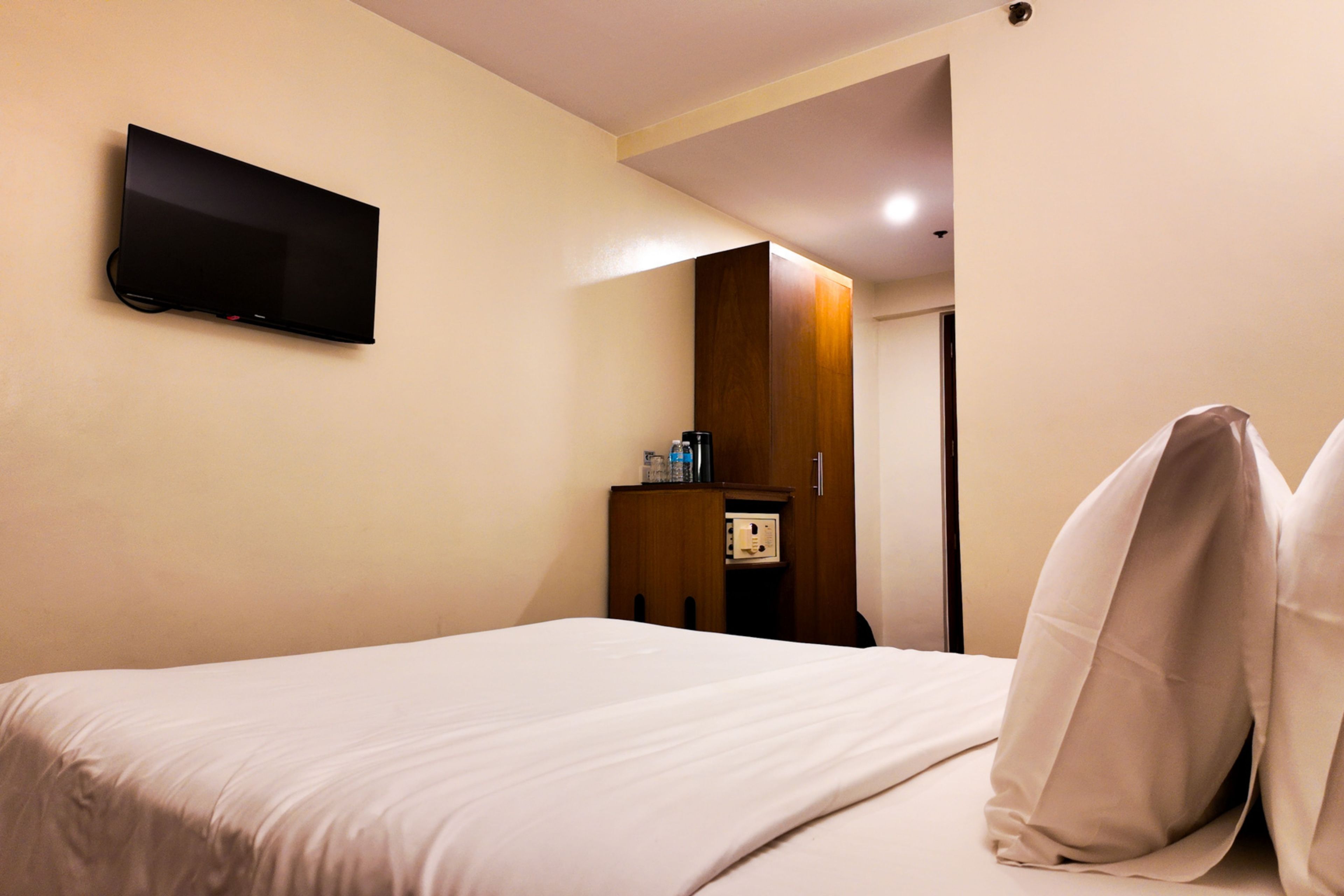 A hotel room with a bed, a flat-screen TV mounted on the wall, a wooden wardrobe, and a small table with a coffee maker.