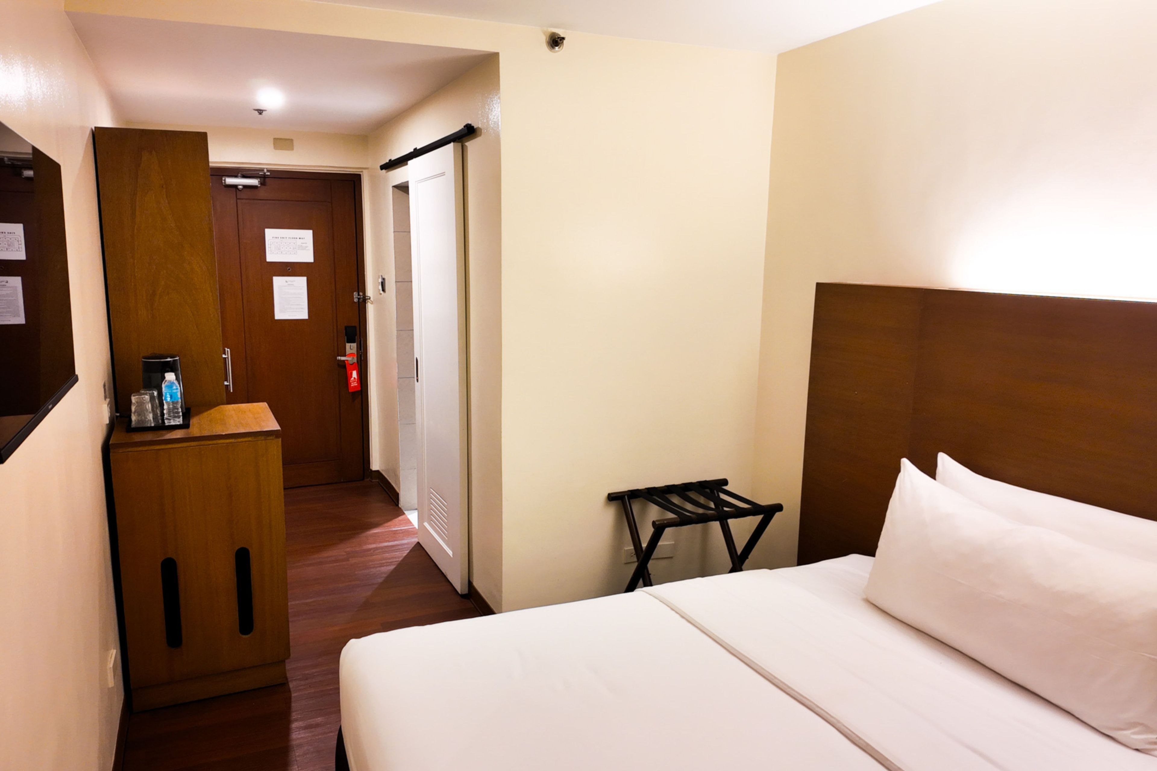 Una habitación de hotel con una cama, un escritorio de madera, una mesita pequeña y una puerta que da a un pasillo.