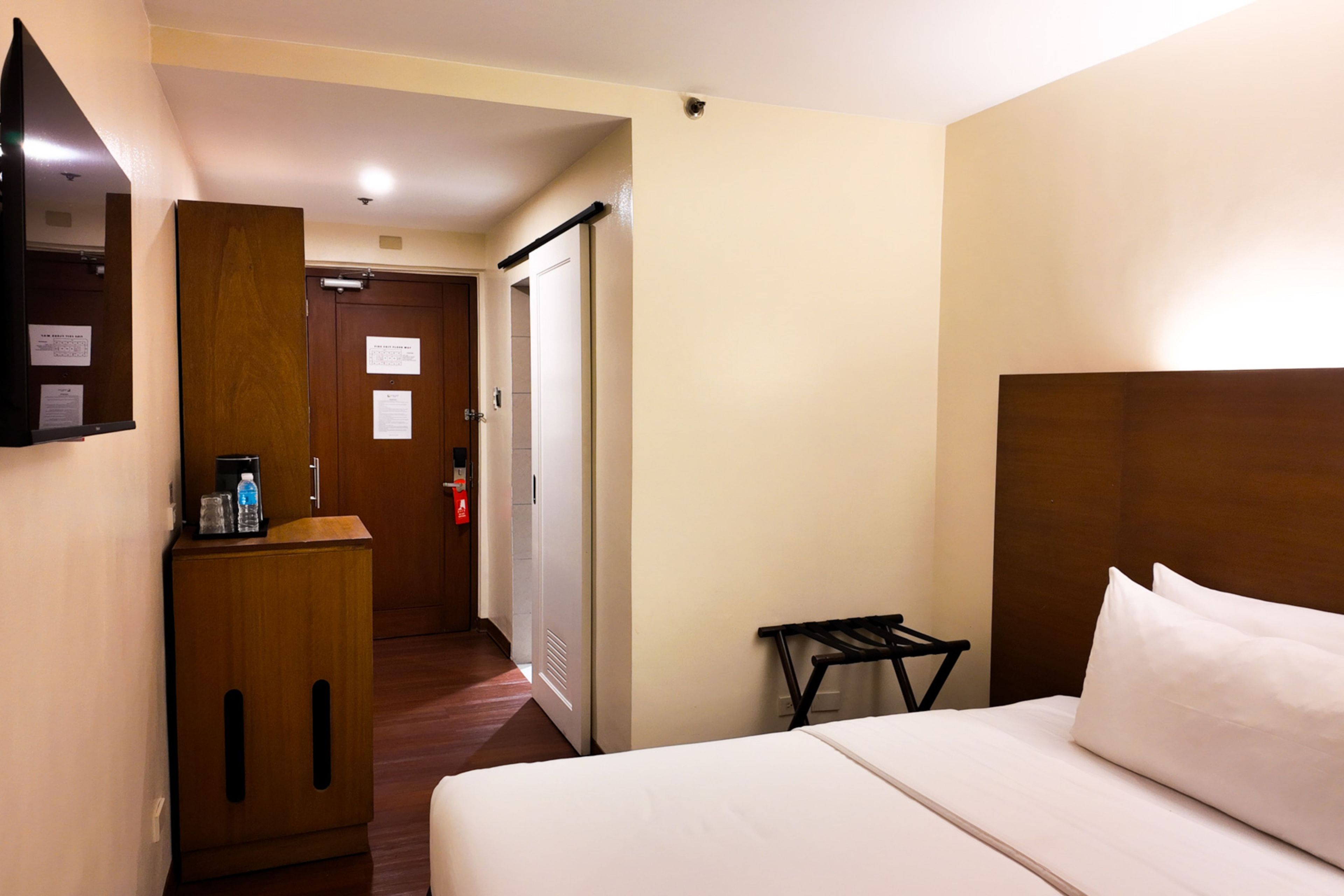 A hotel room with a bed, a desk with a water bottle, a mirror, and a door leading to another room.
